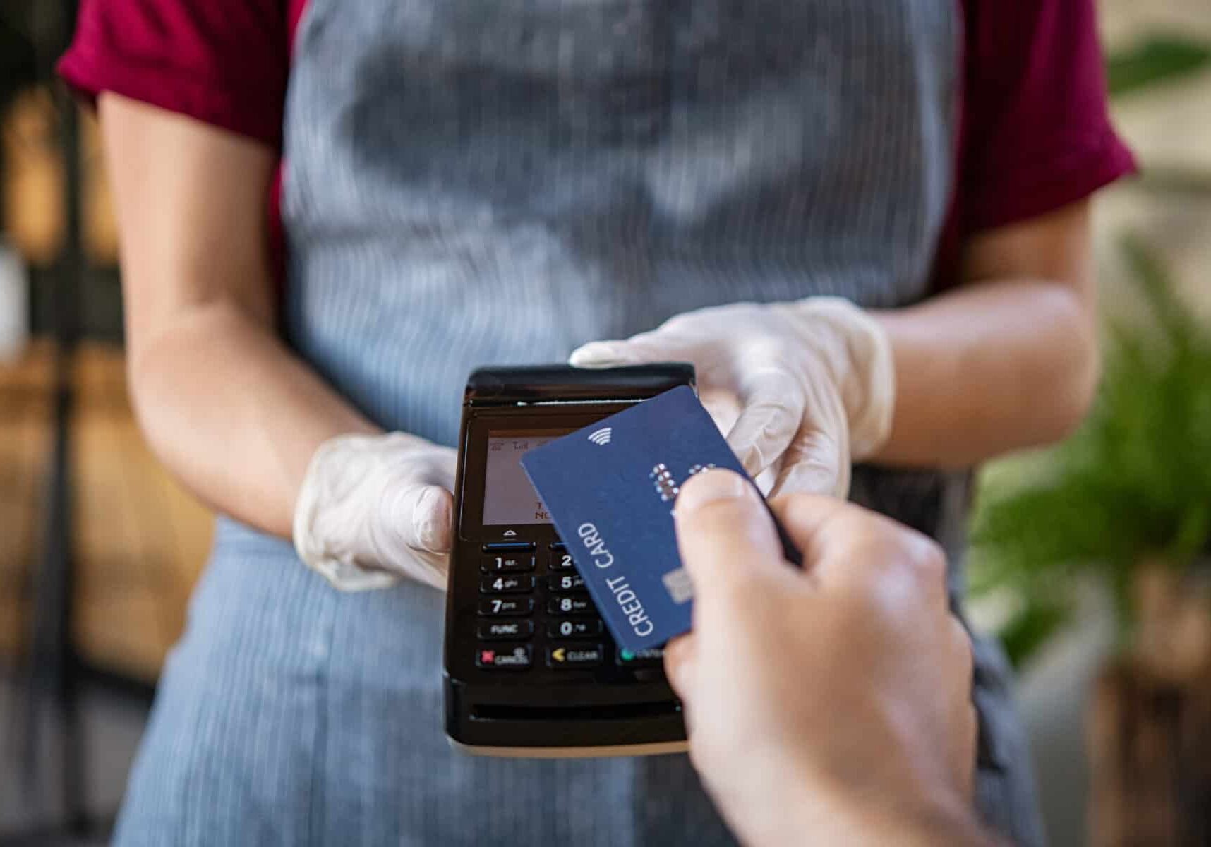 Waitress holding credit card reader machine and wearing protective disposable gloves at bar counter with client holding credit card. Man paying bill with credit card. Hand of customer paying with contactless credit card with NFC technology.