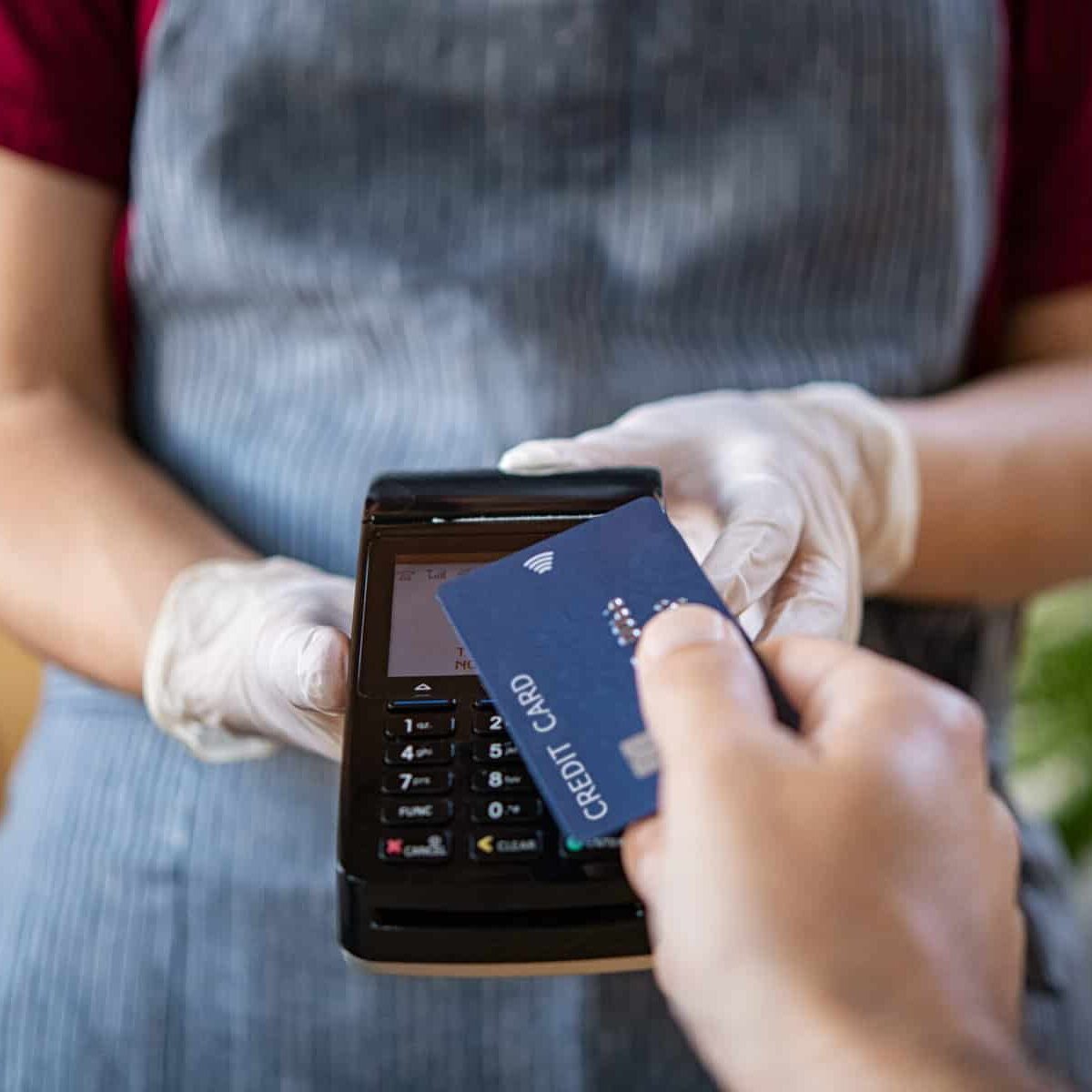 Waitress holding credit card reader machine and wearing protective disposable gloves at bar counter with client holding credit card. Man paying bill with credit card. Hand of customer paying with contactless credit card with NFC technology.